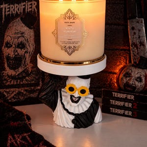 May include: A three-wick White Barn White Pumpkin candle in a frosted glass jar sits on a white pedestal held by a clown figurine. The clown has a black and white outfit, a ruffled collar, and yellow sunflower-shaped glasses on its face.