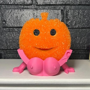 以下が含まれることがあります： 笑顔の顔とピンク色の胸の形をしたベースが付いた、オレンジ色のキラキラ光るカボチャ型のオブジェクト。ベースには腕とピンクのリボンが含まれています。装飾品またはノベルティ製品である可能性があります。