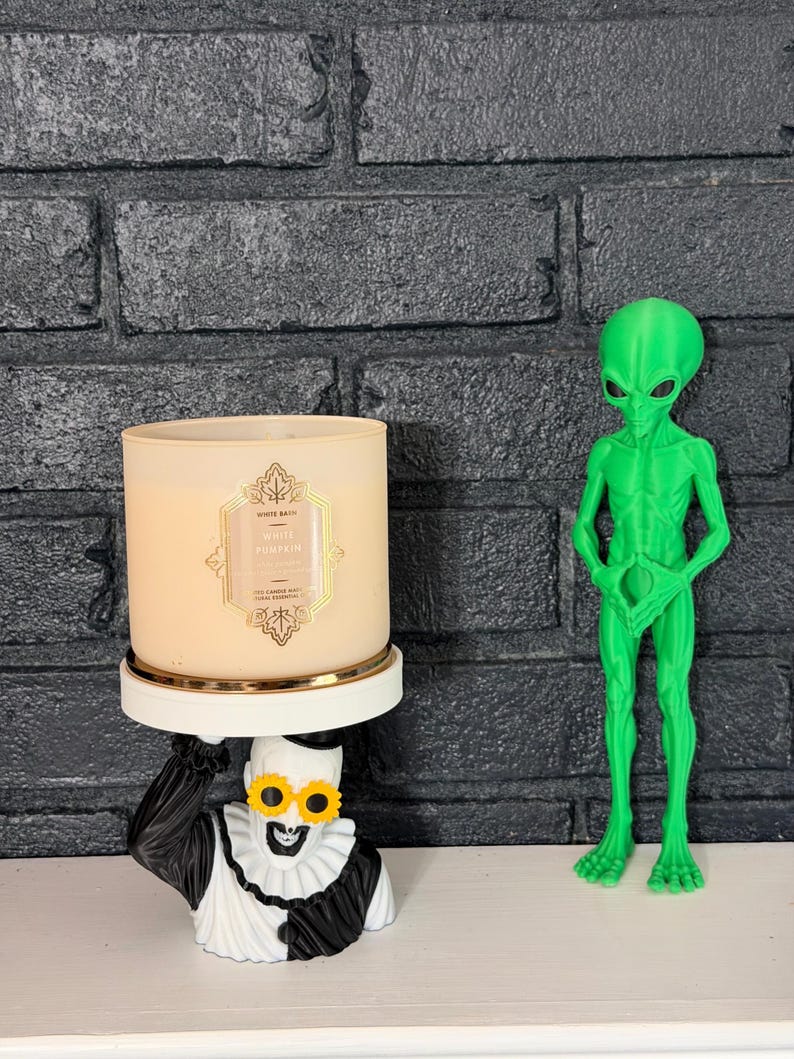 May include: A beige three-wick candle labeled White Pumpkin sits on a white pedestal held by a black and white clown figure wearing yellow flower-shaped glasses. Beside it stands a bright green standing alien figurine with large black eyes.