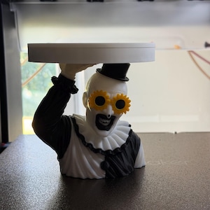 May include: A 3D printed bust of a clown figure wearing a black top hat and yellow sunflower-shaped glasses. The clown has a white face with black makeup, a ruffled collar, and a black and white outfit. The figure holds a flat white circular tray.