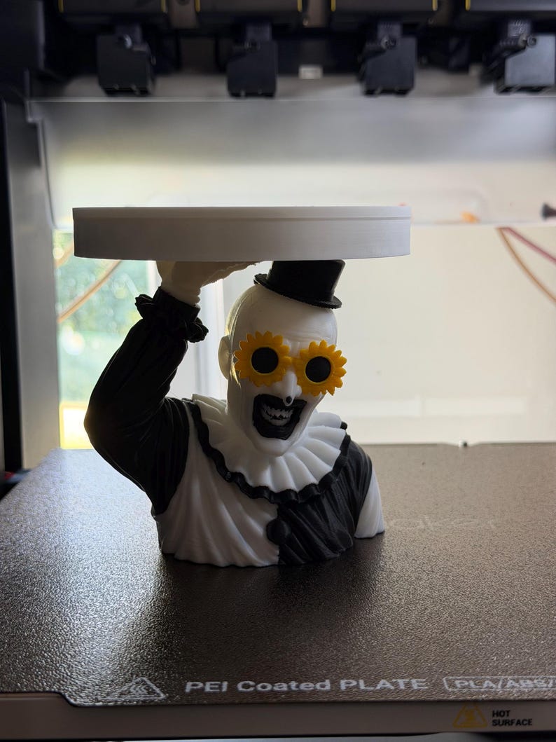 May include: A 3D printed bust of a clown figure wearing a black top hat and yellow sunflower-shaped glasses. The clown has a white face with black makeup, a ruffled collar, and a black and white outfit. The figure holds a flat white circular tray.