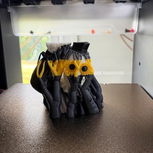 May include: A 3D printed figurine featuring two figures with black and white bodies, wearing black top hats. The figures have yellow sunflower-shaped faces with black centers, and their black arms are raised to cover their faces in a pose.