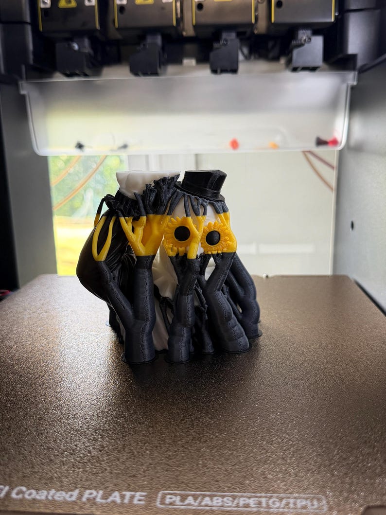 May include: A 3D printed figurine featuring two figures with black and white bodies, wearing black top hats. The figures have yellow sunflower-shaped faces with black centers, and their black arms are raised to cover their faces in a pose.
