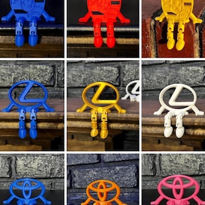 May include: A collection of colorful, articulated figures featuring car brand logos. The figures are in various colors, including blue, red, yellow, gold, white, orange, and pink. Each figure has a logo on its body and jointed legs.