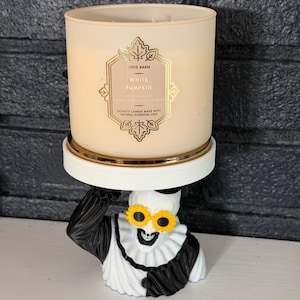 May include: A White Barn White Pumpkin scented candle in a beige glass jar with a gold label, resting on a white pedestal stand supported by a black and white clown figure wearing yellow sunflower-shaped glasses and a ruffled collar.