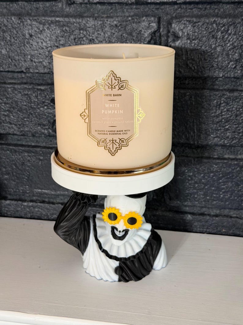 May include: A White Barn White Pumpkin scented candle in a beige glass jar with a gold label, resting on a white pedestal stand supported by a black and white clown figure wearing yellow sunflower-shaped glasses and a ruffled collar.