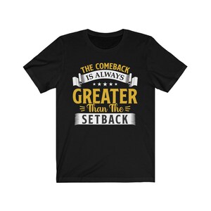 May include: Black t-shirt with the text "THE COMEBACK IS ALWAYS GREATER Than The SETBACK" in white and gold lettering. The design includes stars and banner-style elements. The shirt is a short-sleeved crew neck.