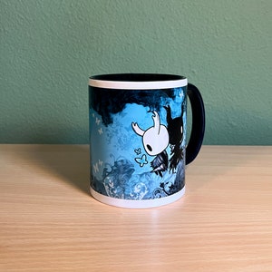 Puede incluir: Una taza de cer&aacute;mica azul y negra con un borde blanco. La taza presenta una ilustraci&oacute;n estilizada de dos personajes del videojuego Hollow Knight. Los personajes est&aacute;n rodeados de un dise&ntilde;o floral azul y blanco.