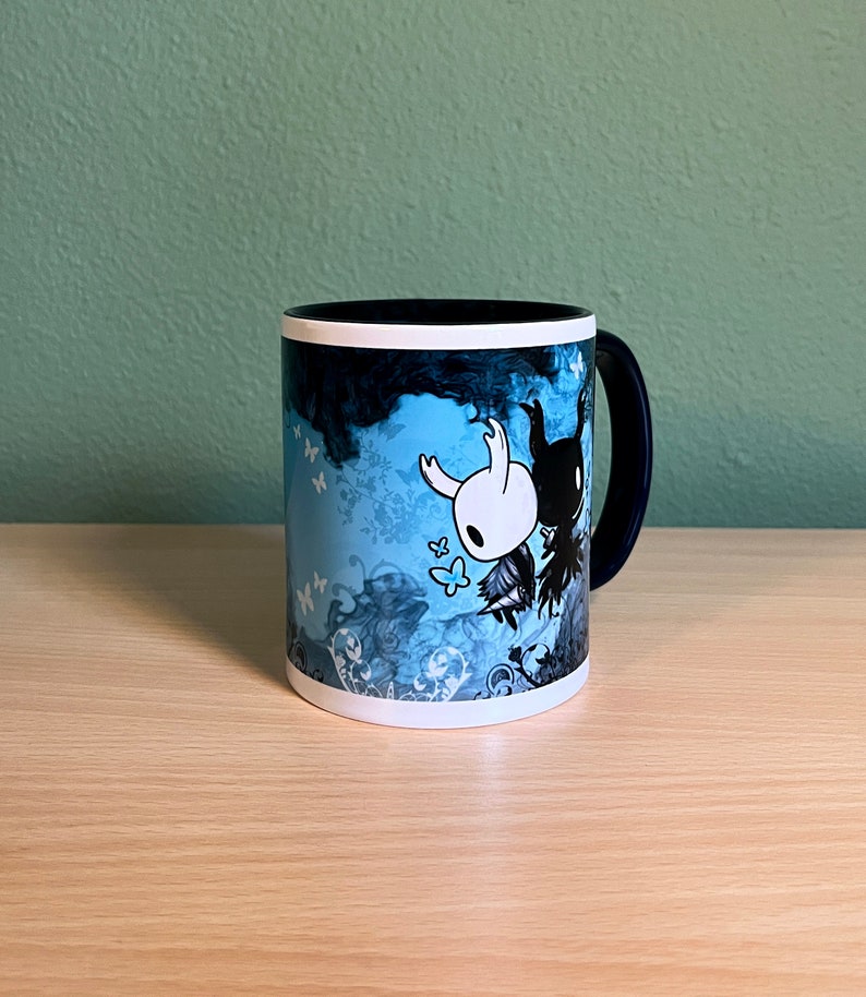 Puede incluir: Una taza de cer&aacute;mica azul y negra con un borde blanco. La taza presenta una ilustraci&oacute;n estilizada de dos personajes del videojuego Hollow Knight. Los personajes est&aacute;n rodeados de un dise&ntilde;o floral azul y blanco.