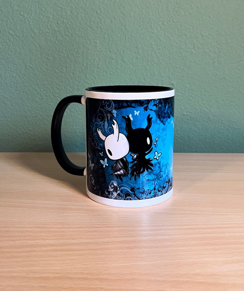 Puede incluir: Una taza de cer&aacute;mica blanca con asa negra. La taza presenta un dise&ntilde;o azul y negro con dos personajes del videojuego Hollow Knight. Los personajes son un caballero blanco y un caballero negro. El fondo es un remolino azul y negro con mariposas.