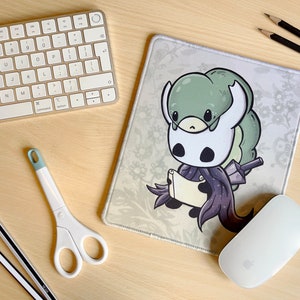 Könnte beinhalten: Eine weiße und grüne Mausmatte mit einer Cartoon-Illustration einer Figur aus dem Videospiel Hollow Knight. Die Figur hält ein Buch und hat einen großen, grünen, insektenartigen Kopf.