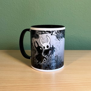 May include: A black and white ceramic mug with a graphic of two characters from the video game Hollow Knight. The characters are a knight and a creature with a long, thin body and a large head. The background is a swirling pattern of black and white.