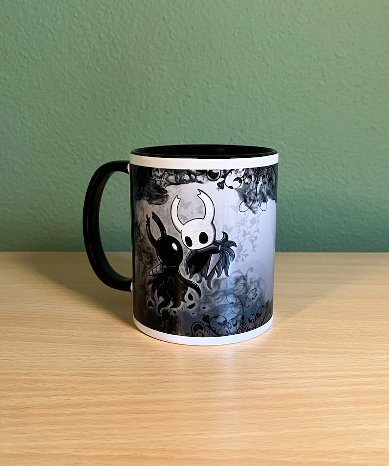 May include: A black and white ceramic mug with a graphic of two characters from the video game Hollow Knight. The characters are a knight and a creature with a long, thin body and a large head. The background is a swirling pattern of black and white.