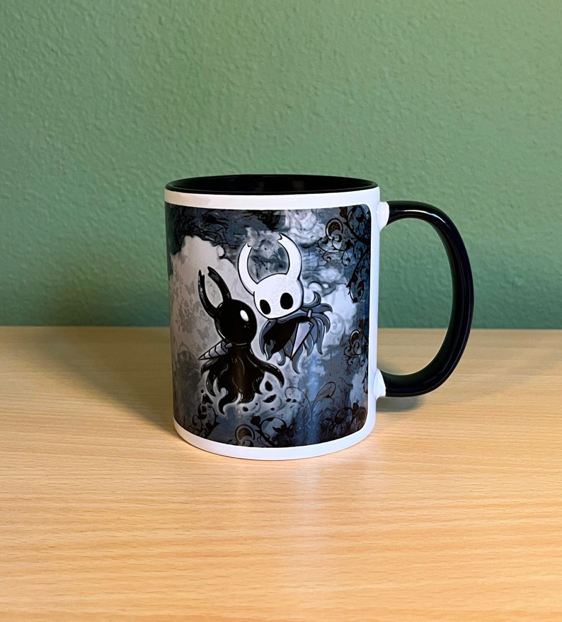 May include: A white ceramic mug with a black handle. The mug features a black and white illustration of two characters from the video game Hollow Knight. The characters are facing each other and appear to be dancing. The background is a dark gray with a floral pattern.