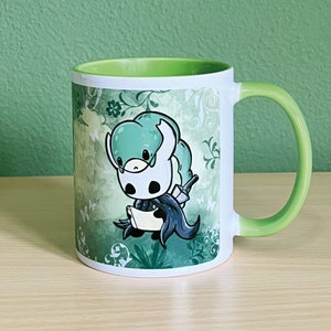 May include: A white ceramic mug with a green rim and handle. The mug features a cartoon illustration of a green and white knight with a sword and shield, set against a green and white floral background.
