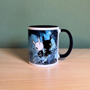 Puede incluir: Una taza de cer&aacute;mica blanca con asa negra. La taza presenta un dise&ntilde;o azul y negro con dos personajes del videojuego Hollow Knight. Los personajes son un caballero blanco y un caballero negro.