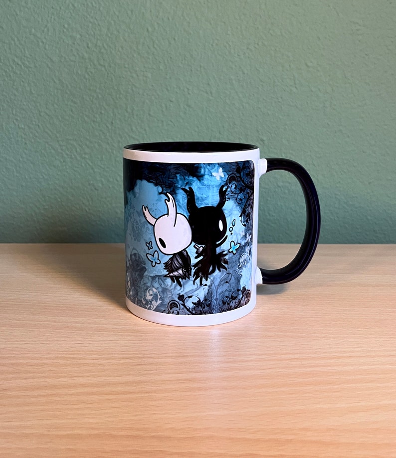 Puede incluir: Una taza de cer&aacute;mica blanca con asa negra. La taza presenta un dise&ntilde;o azul y negro con dos personajes del videojuego Hollow Knight. Los personajes son un caballero blanco y un caballero negro.