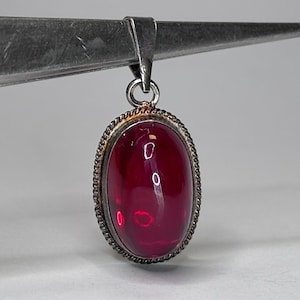 May include: An oval pendant featuring a smooth, deep red cabochon stone set in a silver-toned metal frame with a twisted rope-style border. The pendant includes a triangular bail at the top, suspended from a pair of metal tweezers.