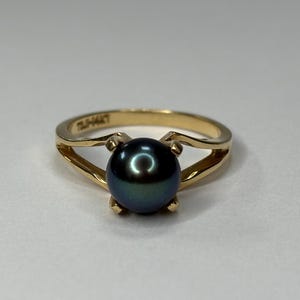 Puede incluir: Un anillo de oro con una perla oscura e iridiscente. El anillo tiene un dise&ntilde;o de ca&ntilde;a dividida, con la perla sujeta por cuatro garras. El anillo est&aacute; sobre un fondo neutro.