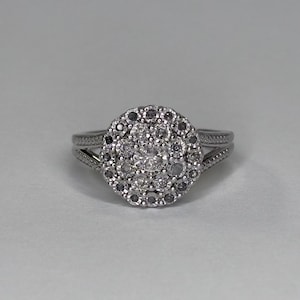 May include: A silver-toned ring featuring a circular cluster of small, clear, round-cut stones arranged in a floral pattern. The central stone is surrounded by two concentric rings of smaller stones, set on a split-shank band with textured detailing.