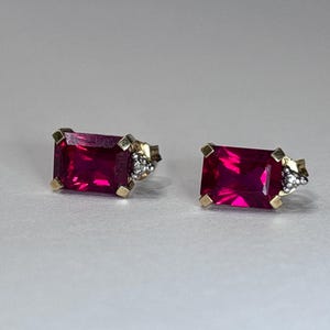 May include: A pair of gold-toned stud earrings, each featuring a rectangular, vibrant magenta gemstone. The earrings have small, decorative accents with clear stones on the side.