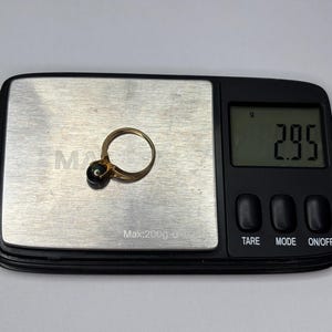 Puede incluir: Un anillo de oro con una piedra oscura se coloca en una balanza digital. La balanza muestra un peso de 2,95 gramos. El anillo est&aacute; centrado en la superficie met&aacute;lica de la balanza, con la pantalla digital al fondo.