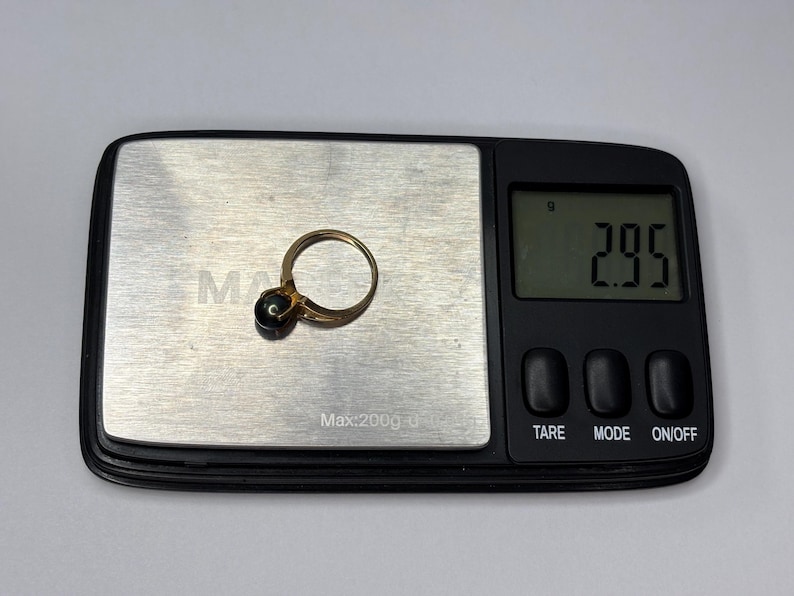 Puede incluir: Un anillo de oro con una piedra oscura se coloca en una balanza digital. La balanza muestra un peso de 2,95 gramos. El anillo est&aacute; centrado en la superficie met&aacute;lica de la balanza, con la pantalla digital al fondo.