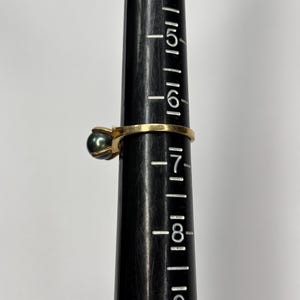 Puede incluir: Un anillo de oro con una perla gris oscuro. El anillo est&aacute; en un medidor de anillos negro. El medidor de anillos tiene medidas de 4 a 9. Una joya cl&aacute;sica.