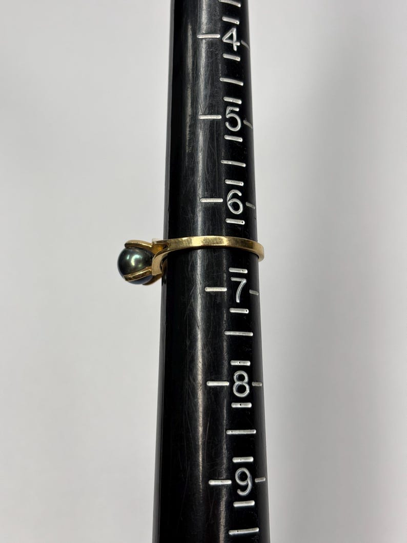 Puede incluir: Un anillo de oro con una perla gris oscuro. El anillo est&aacute; en un medidor de anillos negro. El medidor de anillos tiene medidas de 4 a 9. Una joya cl&aacute;sica.