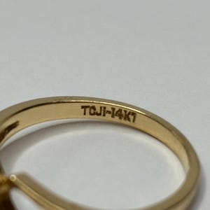 Puede incluir: Primer plano de un anillo de oro con una piedra preciosa oscura. La banda del anillo est&aacute; estampada con "TCJI-14K1". El anillo est&aacute; sobre un fondo blanco. El anillo es probablemente una joya.