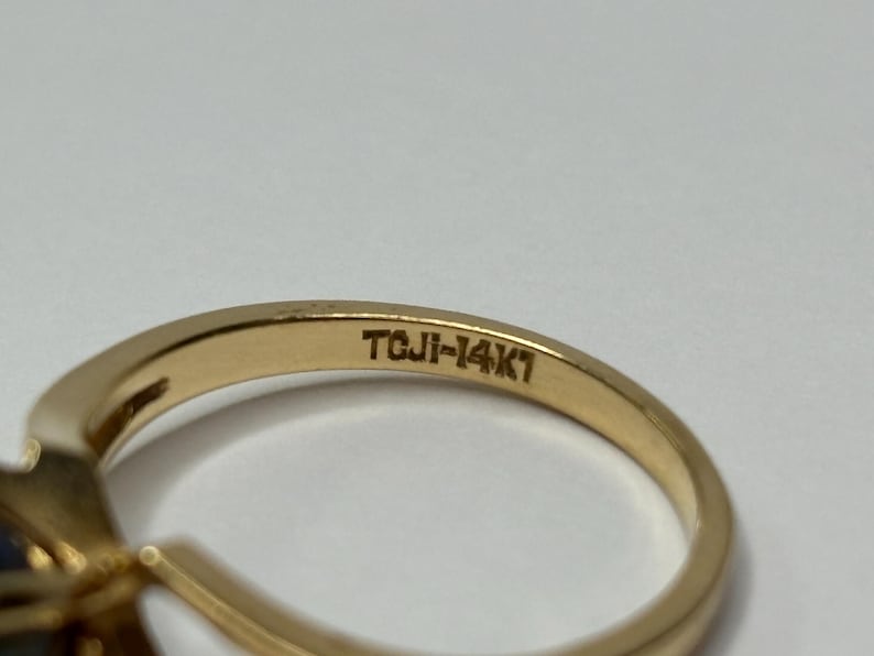 Puede incluir: Primer plano de un anillo de oro con una piedra preciosa oscura. La banda del anillo est&aacute; estampada con "TCJI-14K1". El anillo est&aacute; sobre un fondo blanco. El anillo es probablemente una joya.