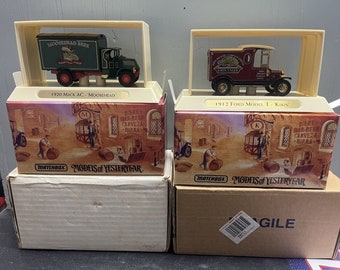 May include: Two vintage toy trucks in display boxes. The trucks are green and red and feature the logos of Moosehead Beer and Kirin Lager. The boxes are brown with a vintage-style illustration of a brewery.