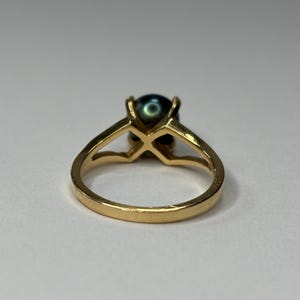 Puede incluir: Un anillo de oro con una perla oscura e iridiscente. El anillo tiene un dise&ntilde;o solitario cl&aacute;sico con una ca&ntilde;a dividida y un engaste de cuatro puntas. La banda es de color oro c&aacute;lido y la perla tiene un brillo profundo y lustroso.