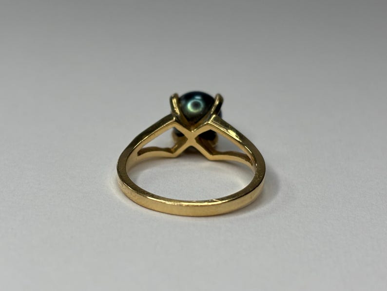 Puede incluir: Un anillo de oro con una perla oscura e iridiscente. El anillo tiene un dise&ntilde;o solitario cl&aacute;sico con una ca&ntilde;a dividida y un engaste de cuatro puntas. La banda es de color oro c&aacute;lido y la perla tiene un brillo profundo y lustroso.