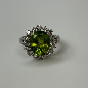 Puede incluir: Un anillo de plata con una gran piedra preciosa ovalada verde rodeada por un halo de diamantes más pequeños.