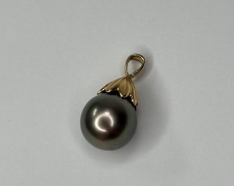 Puede incluir: Un colgante de perla gris oscuro con una fianza y un engaste decorativo dorados. La perla tiene una superficie lisa y reflectante. El colgante está diseñado para ser usado como collar.