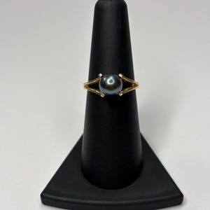 Puede incluir: Un anillo de oro con una perla oscura e iridiscente. El anillo tiene un dise&ntilde;o de banda dividida, con la perla sujeta por dos puntas de oro. El anillo se exhibe en un soporte negro para anillos sobre un fondo blanco.