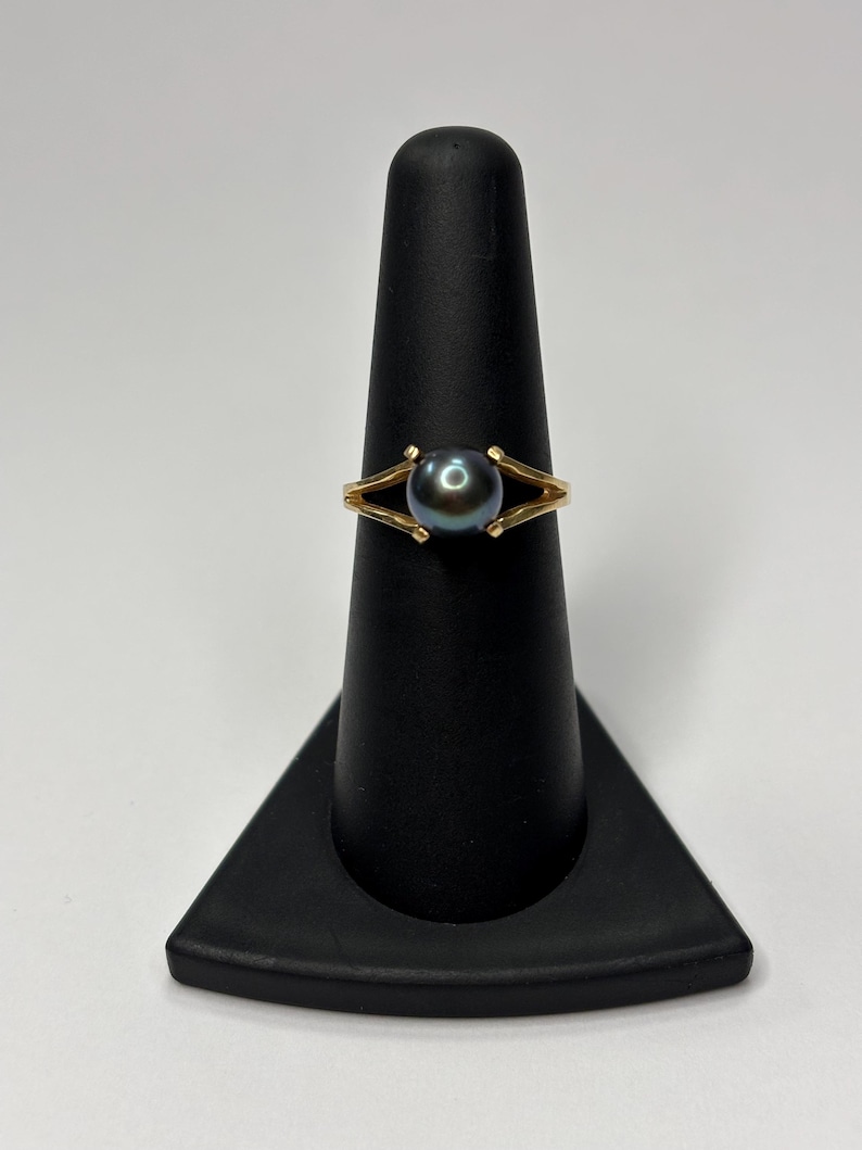 Puede incluir: Un anillo de oro con una perla oscura e iridiscente. El anillo tiene un dise&ntilde;o de banda dividida, con la perla sujeta por dos puntas de oro. El anillo se exhibe en un soporte negro para anillos sobre un fondo blanco.