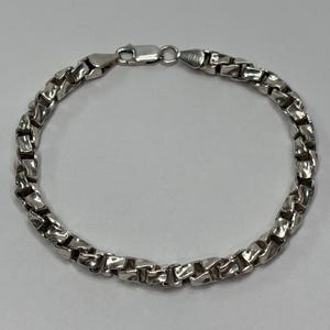 Könnte beinhalten: Ein silbernes Armband mit einem Box-Chain-Design. Das Armband besteht aus glänzendem, metallischem Material und verfügt über einen sicheren Verschluss. Ein stilvolles Accessoire.