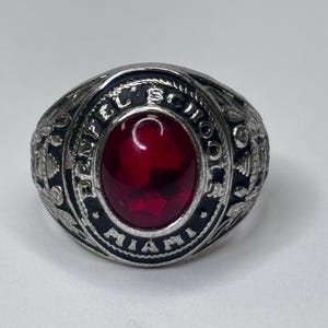 May include: Silver-toned ring with a large, oval, red gemstone at the center. The ring is engraved with the words "BENFEL SCHOOL MIAMI" around the gemstone. The ring has a detailed, textured design.