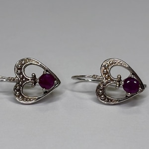 May include: A pair of silver-toned earrings featuring a heart-shaped frame with a beaded texture. Each earring contains a single round, deep pink gemstone set in the center of the heart, attached to a curved wire hook for secure placement.