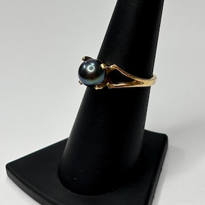 Puede incluir: Un anillo de oro con una perla oscura e iridiscente. La banda del anillo tiene un dise&ntilde;o dividido, y la perla se sujeta con cuatro garras. El anillo se exhibe en un soporte negro para anillos sobre un fondo blanco.