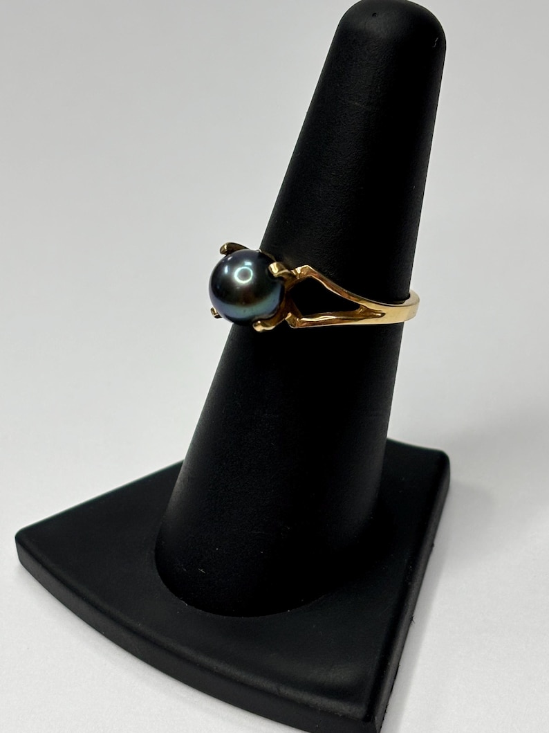 Puede incluir: Un anillo de oro con una perla oscura e iridiscente. La banda del anillo tiene un dise&ntilde;o dividido, y la perla se sujeta con cuatro garras. El anillo se exhibe en un soporte negro para anillos sobre un fondo blanco.