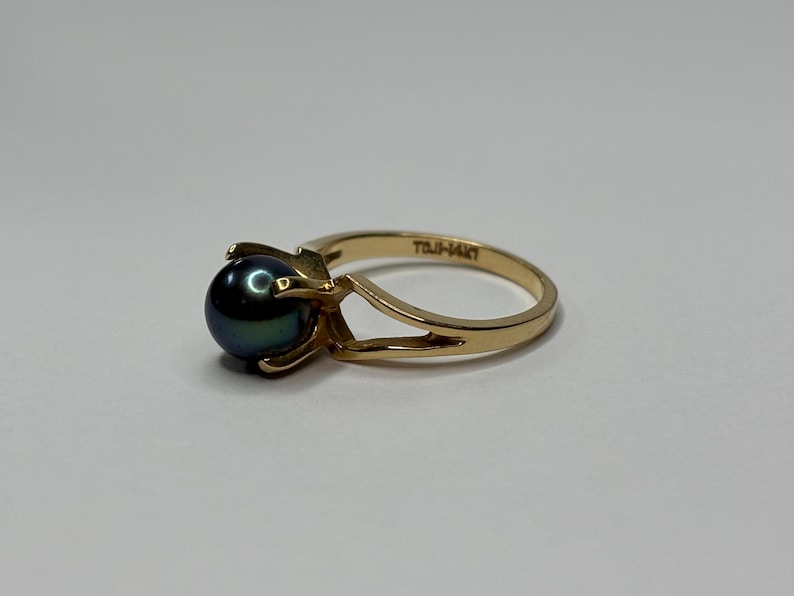 Puede incluir: Un anillo de oro con una perla oscura e iridiscente. El anillo tiene un dise&ntilde;o de ca&ntilde;a dividida y un engaste de cuatro puntas. La banda est&aacute; estampada con "TOJI-14KT".