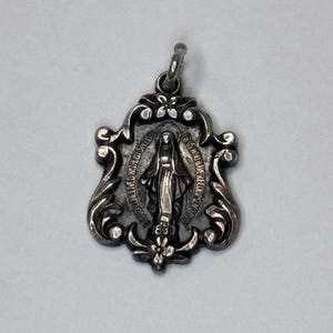May include: A silver-toned pendant with an ornate, shield-shaped frame. The pendant features a central image of a robed figure and surrounding text. The design includes floral accents and a loop for a chain.