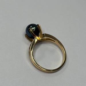 Puede incluir: Un anillo de oro con una perla oscura e iridiscente. La perla est&aacute; engastada en un engaste de cuatro puntas, y la banda es de oro pulido. El anillo se muestra sobre un fondo blanco liso.