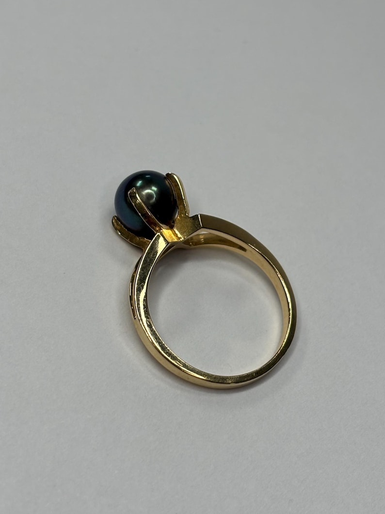 Puede incluir: Un anillo de oro con una perla oscura e iridiscente. La perla est&aacute; engastada en un engaste de cuatro puntas, y la banda es de oro pulido. El anillo se muestra sobre un fondo blanco liso.