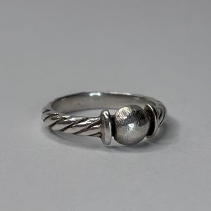 Puede incluir: Un anillo de plata con una banda texturizada y un diseño esférico central. El anillo tiene un patrón retorcido en forma de cuerda en la banda, que conduce a una esfera central flanqueada por dos elementos redondeados más pequeños.