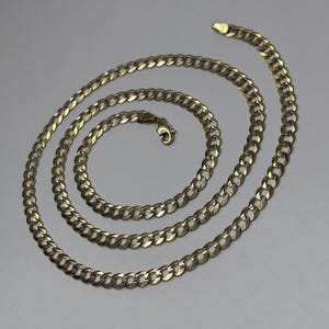 Puede incluir: Un collar de cadena de oro, dispuesto en espiral. La cadena está compuesta por eslabones ovalados entrelazados, con un cierre de mosquetón. El collar se presenta sobre un fondo neutro.