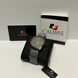 以下が含まれることがあります： グレーのナイロンストラップと白い文字盤が付いたグレーの腕時計。時計はシルバーケースで、文字盤には赤と白のロゴがあります。時計は、黒と白のタグに「Calibre」と書かれた黒いディスプレイスタンドの上に置かれています。